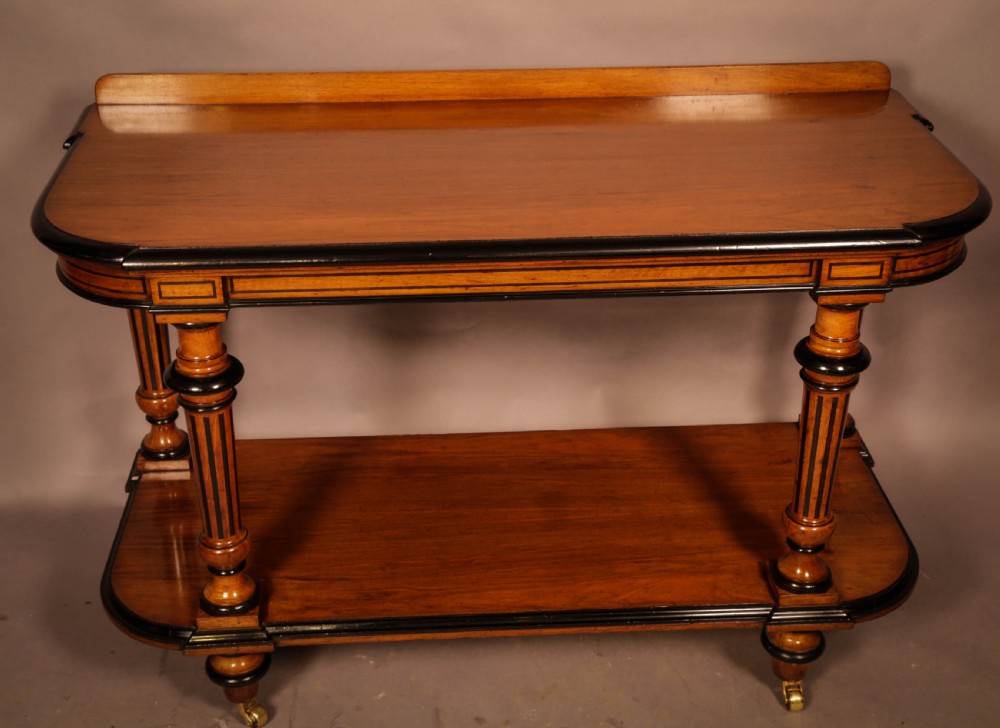 An Aesthetic Movement walnut serving table featuring a rectangular top with a low gallery, ebonised trim, a full lower tier, and turned fluted legs on brass castors.