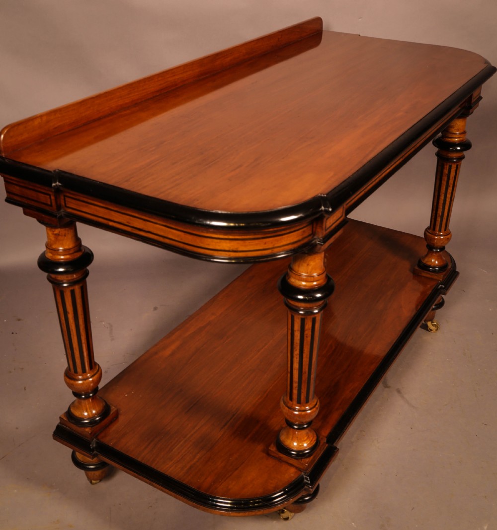 An Aesthetic Movement walnut serving table featuring a rectangular top with a low gallery, ebonised trim, a full lower tier, and turned fluted legs on brass castors. View II