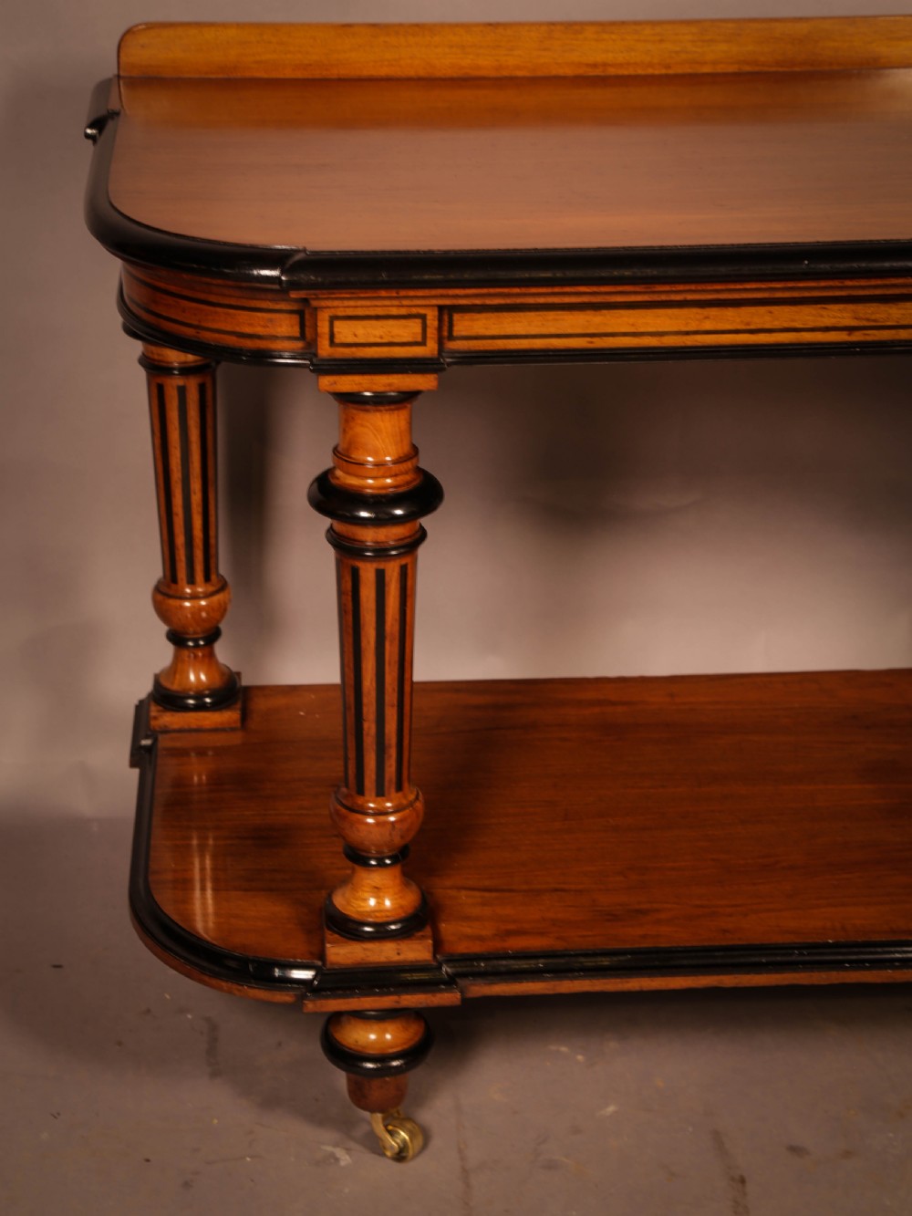 An Aesthetic Movement walnut serving table featuring a rectangular top with a low gallery, ebonised trim, a full lower tier, and turned fluted legs on brass castors. View IV