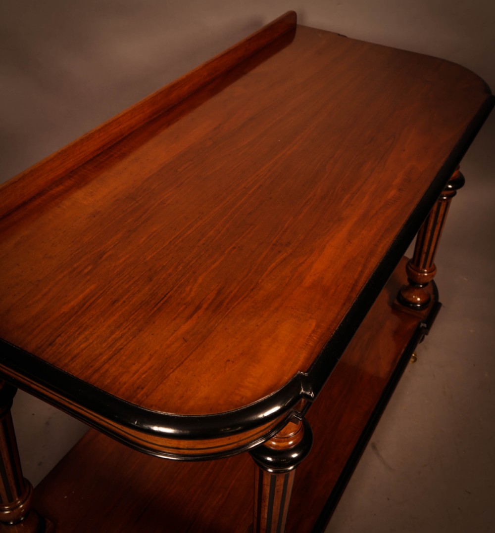 An Aesthetic Movement walnut serving table featuring a rectangular top with a low gallery, ebonised trim, a full lower tier, and turned fluted legs on brass castors. View V