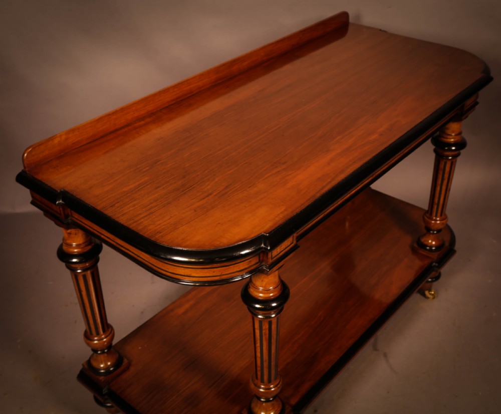 An Aesthetic Movement walnut serving table featuring a rectangular top with a low gallery, ebonised trim, a full lower tier, and turned fluted legs on brass castors. View VI