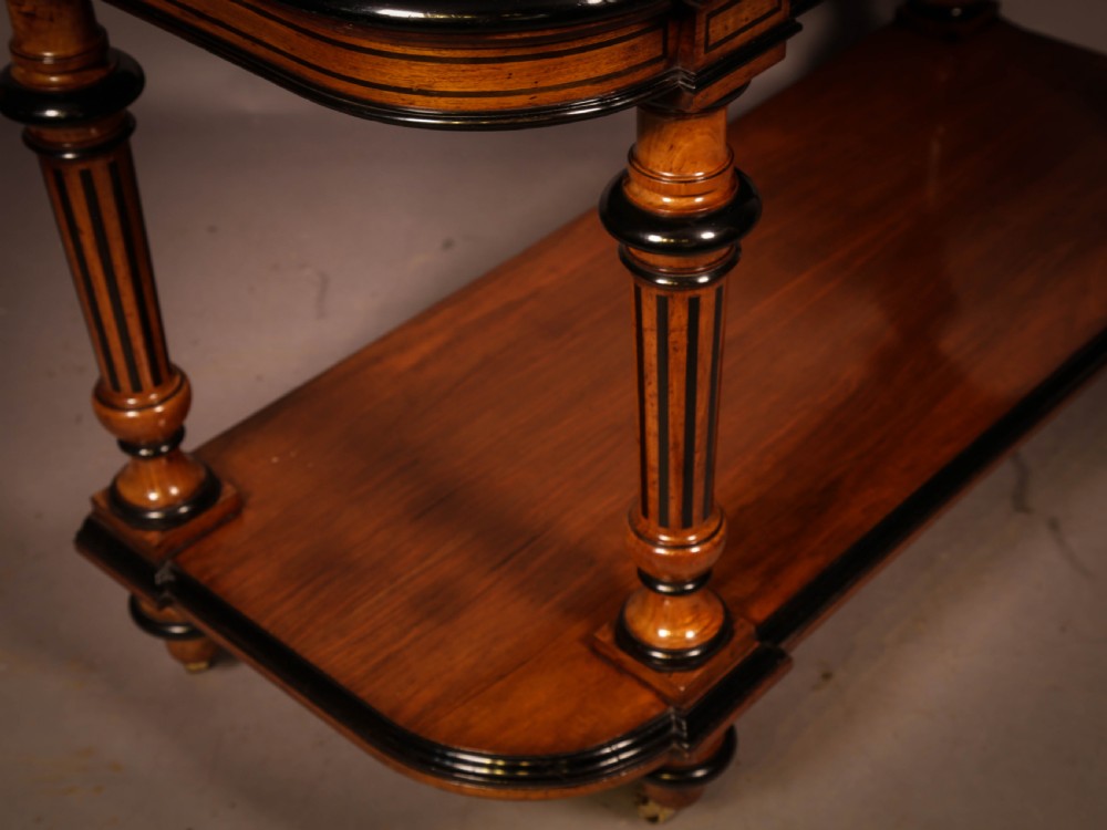 An Aesthetic Movement walnut serving table featuring a rectangular top with a low gallery, ebonised trim, a full lower tier, and turned fluted legs on brass castors. View VII