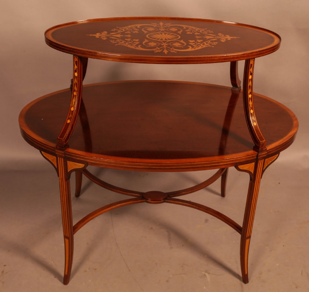 An Edwardian two-tier oval mahogany etagere featuring detailed central floral marquetry inlay, slender curved supports, and swept legs with crossbanding.