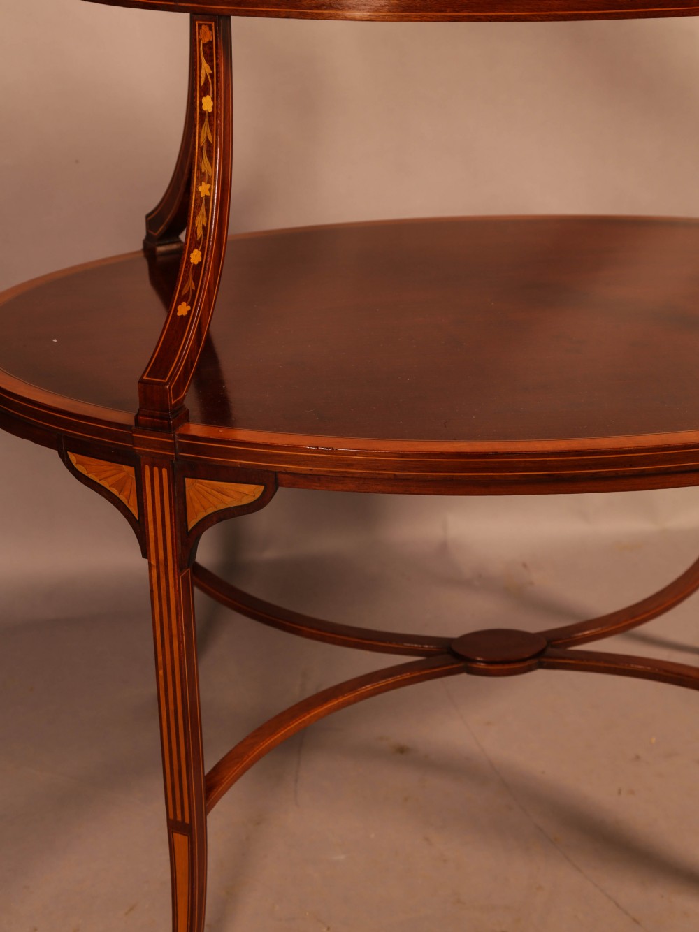 An Edwardian two-tier oval mahogany etagere featuring detailed central floral marquetry inlay, slender curved supports, and swept legs with crossbanding. View X