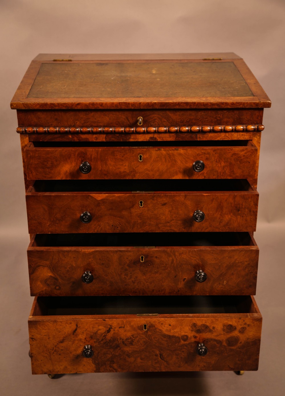 A Victorian burr oak Davenport desk featuring a green leather writing slope, side drawers with wooden knobs, bead moulding, and brass casters. View IV