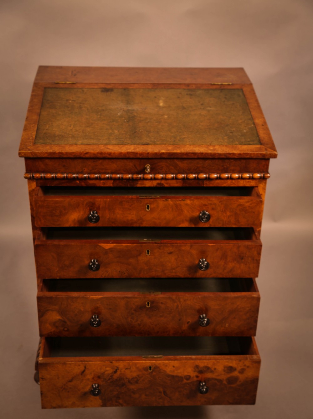 A Victorian burr oak Davenport desk featuring a green leather writing slope, side drawers with wooden knobs, bead moulding, and brass casters. View V