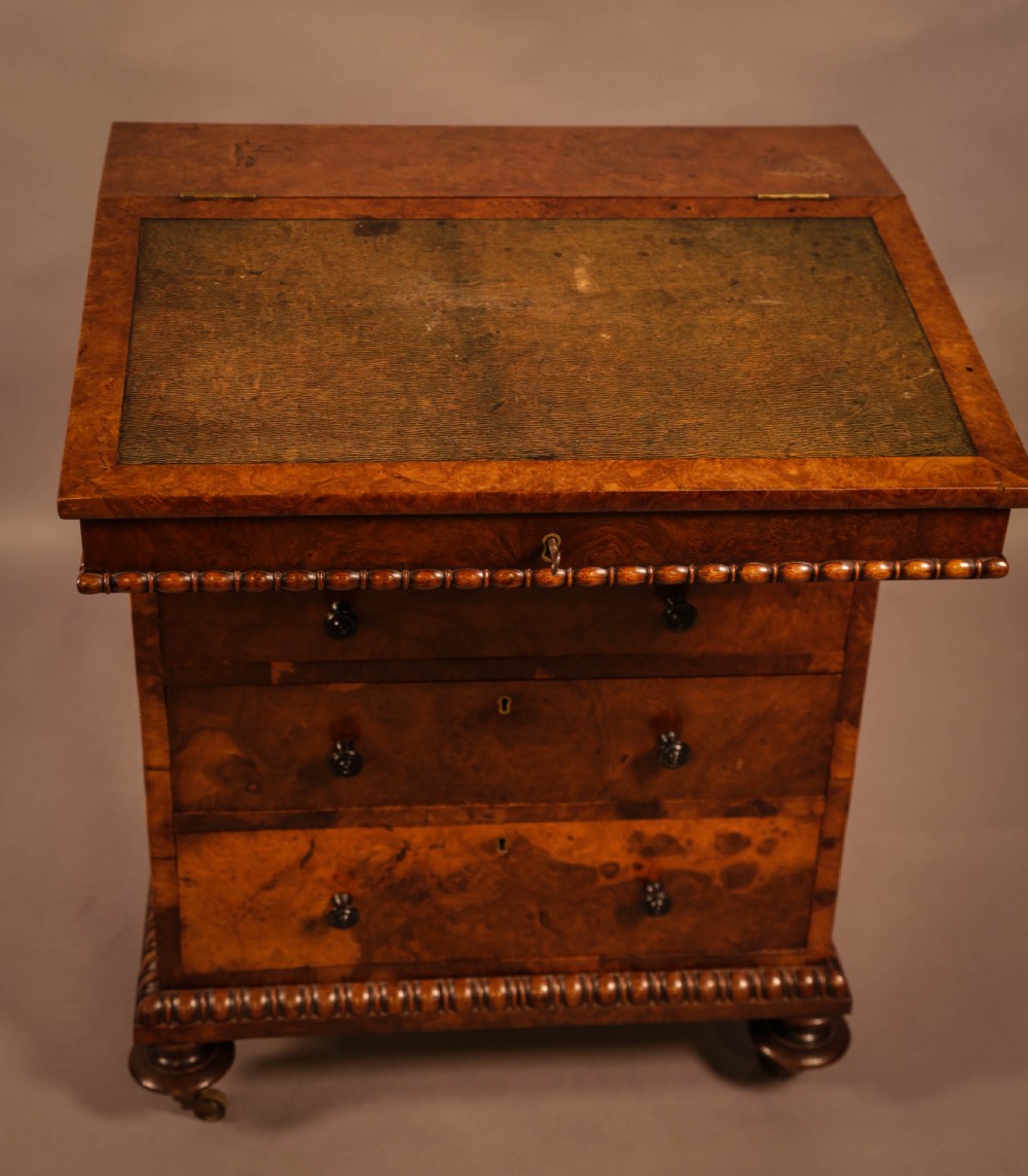 A Victorian burr oak Davenport desk featuring a green leather writing slope, side drawers with wooden knobs, bead moulding, and brass casters. View VI