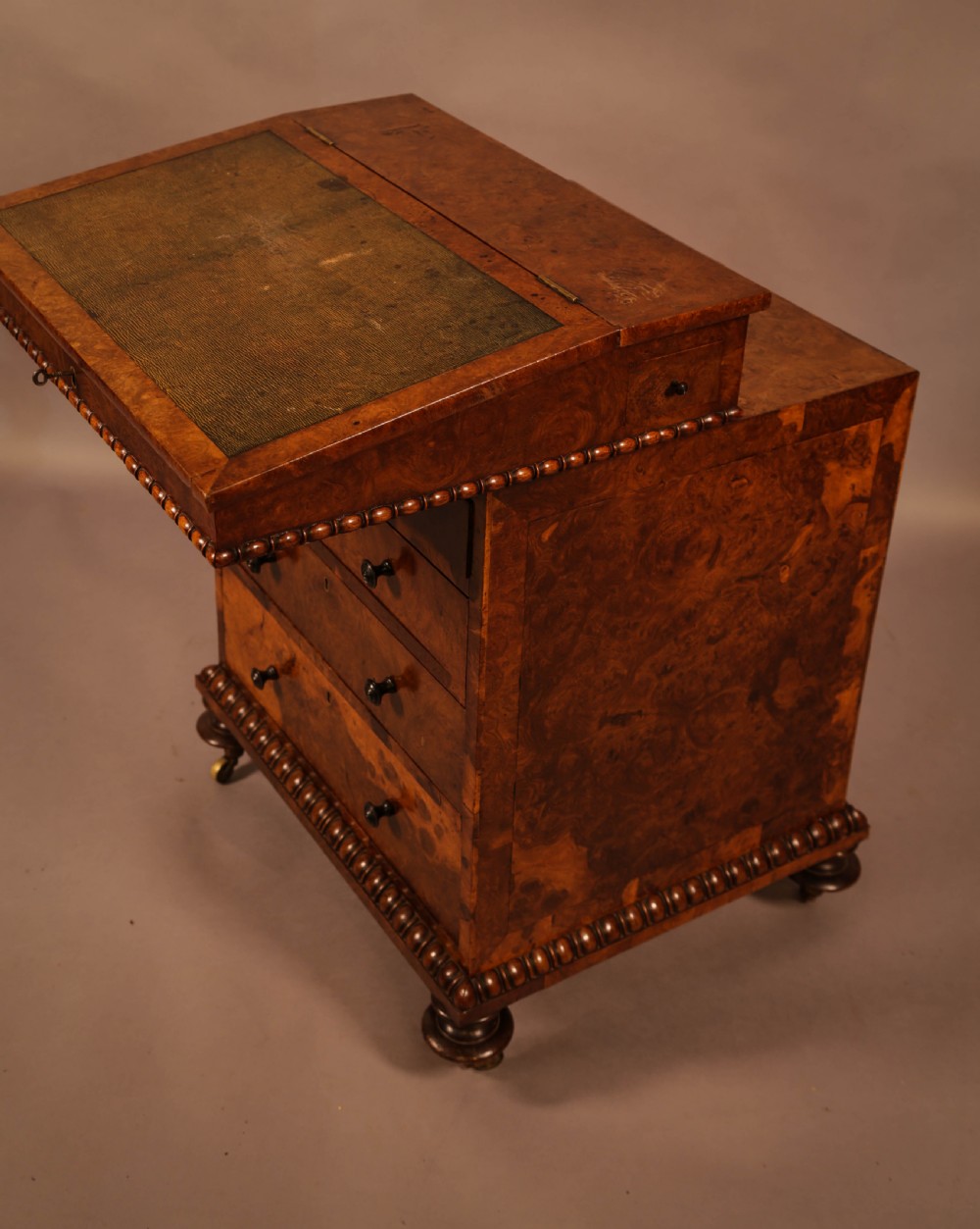 A Victorian burr oak Davenport desk featuring a green leather writing slope, side drawers with wooden knobs, bead moulding, and brass casters. View VII