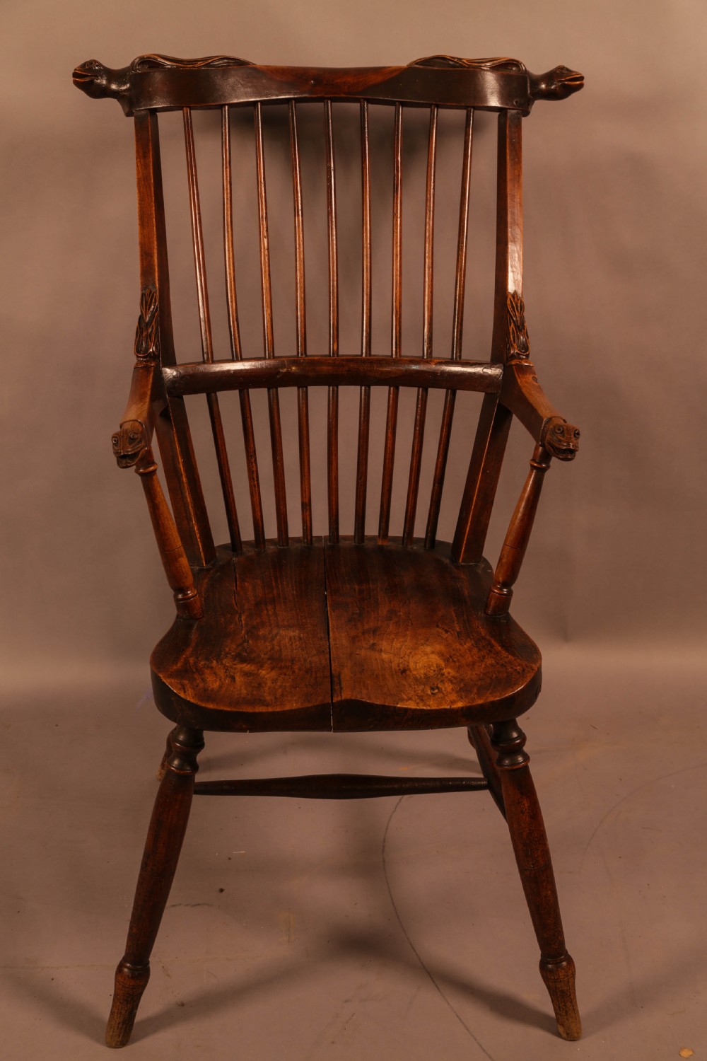 A dark brown antique wooden Windsor armchair featuring vertical spindles, a contoured saddle seat, and hand-carved snake head terminals on the arms and top rail.