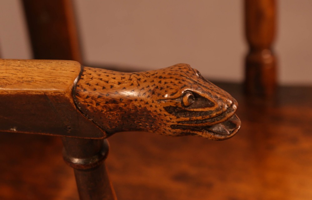 A dark brown antique wooden Windsor armchair featuring vertical spindles, a contoured saddle seat, and hand-carved snake head terminals on the arms and top rail. View V