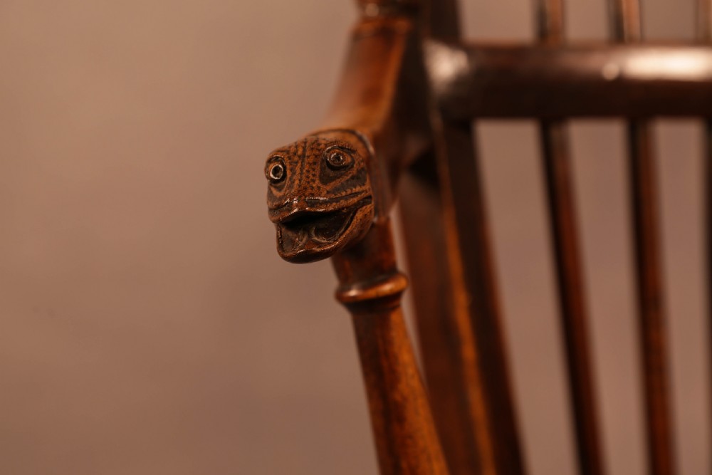 A dark brown antique wooden Windsor armchair featuring vertical spindles, a contoured saddle seat, and hand-carved snake head terminals on the arms and top rail. View XVII