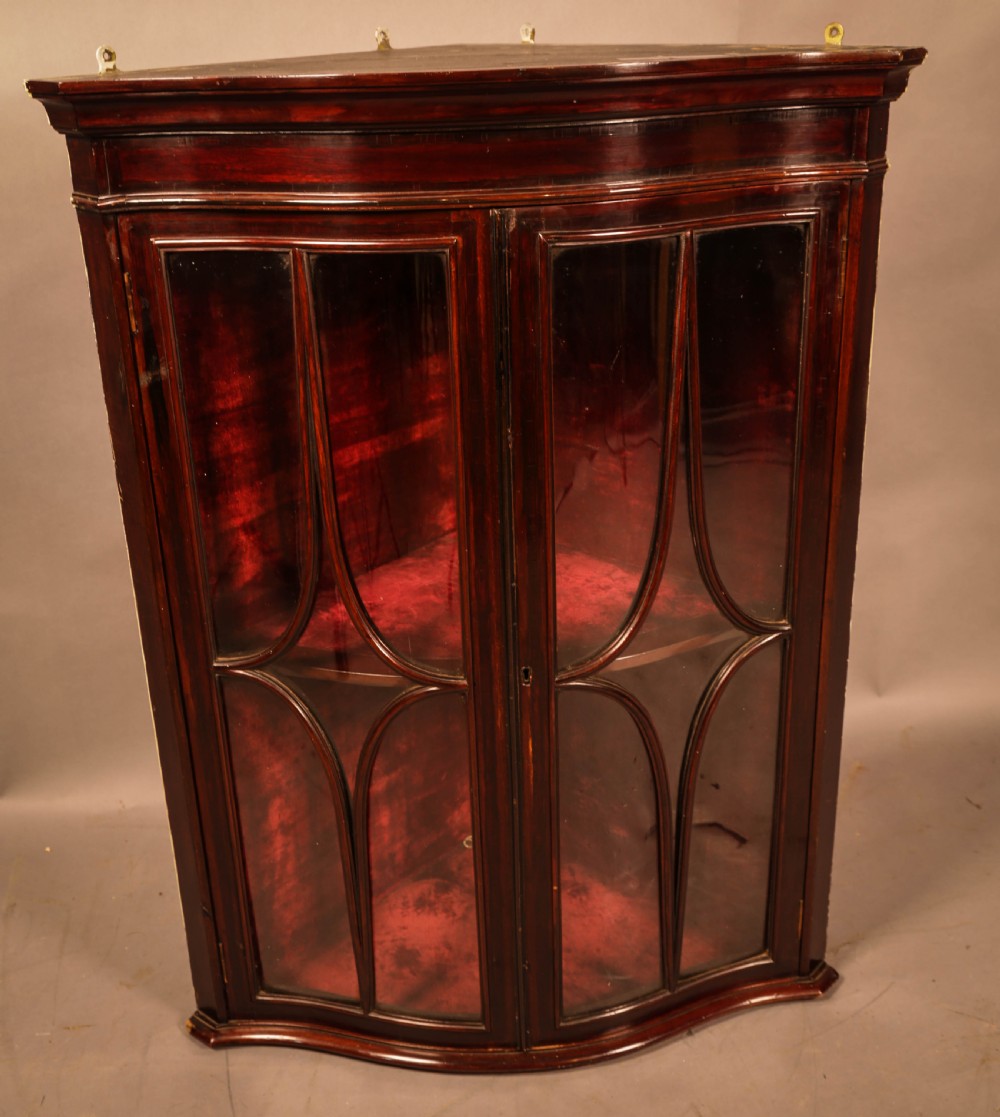 A dark mahogany wall-hanging corner cupboard featuring glazed double doors with serpentine curved fronts and decorative astragal glazing bars.