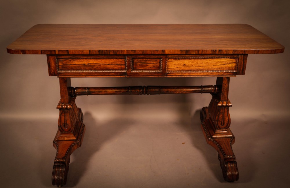 A William IV rosewood library table featuring a rectangular top with rounded corners, frieze drawers, a turned central stretcher, and carved pedestal legs.