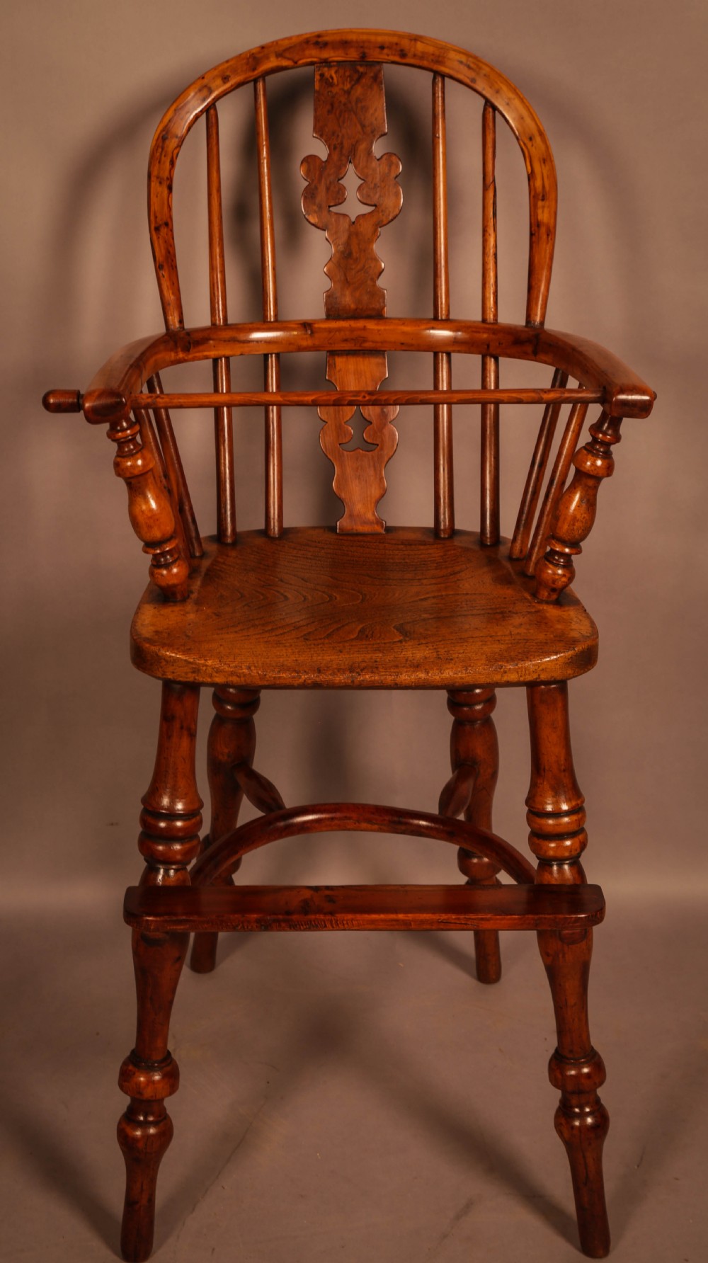 A yew wood Windsor childs high chair with a pierced splat back, turned legs, footrest, and safety bar.