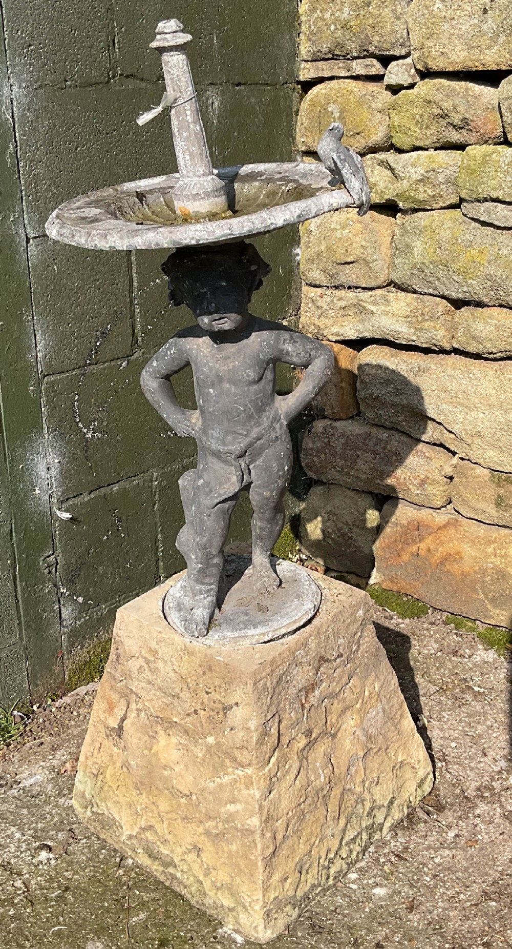 A cast lead fountain featuring a figural base supporting a shallow basin and central spout, mounted upon a rough-hewn stone plinth.