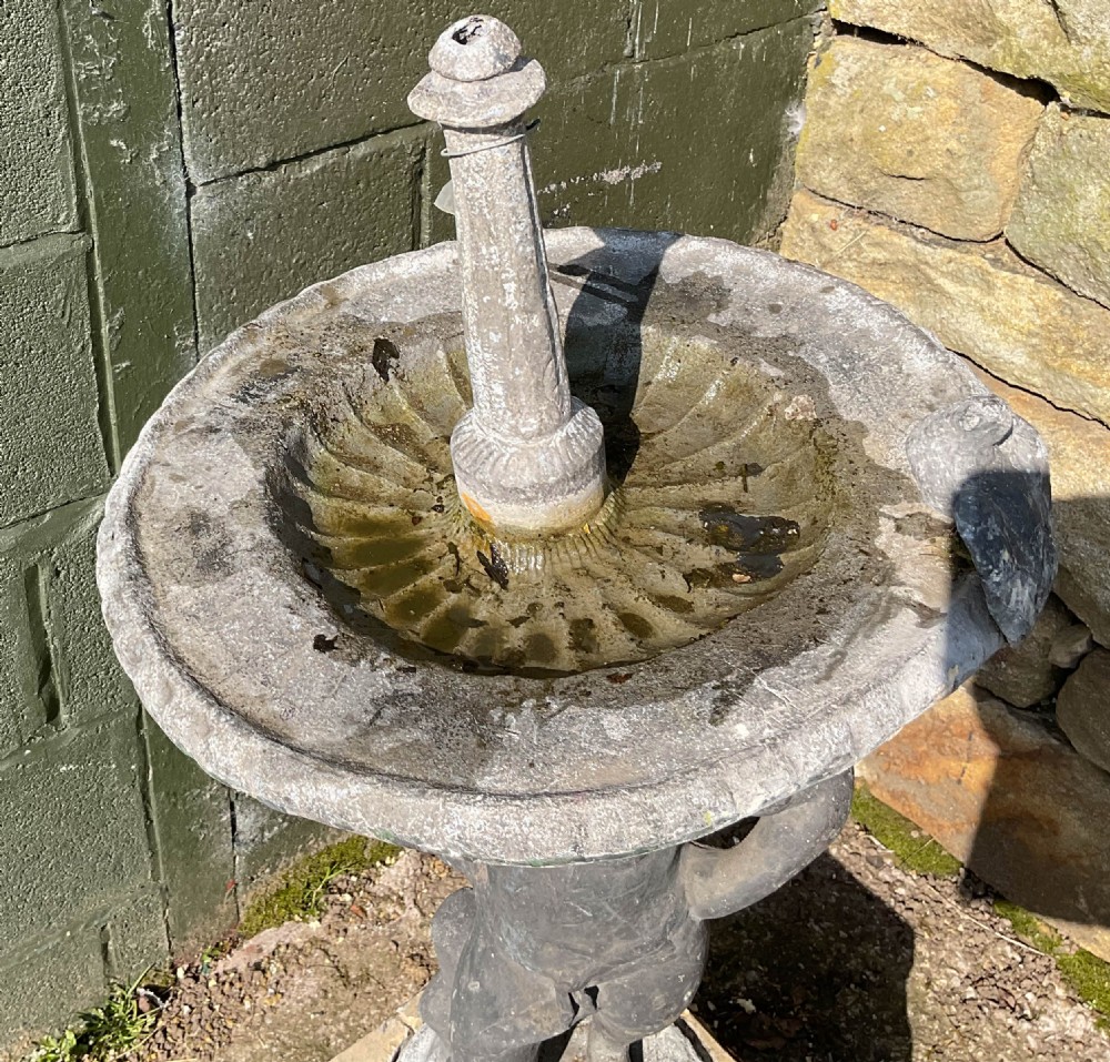 A cast lead fountain featuring a figural base supporting a shallow basin and central spout, mounted upon a rough-hewn stone plinth. View III