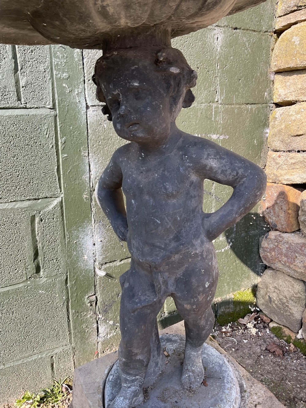 A cast lead fountain featuring a figural base supporting a shallow basin and central spout, mounted upon a rough-hewn stone plinth. View V