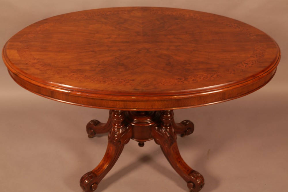 An oval Victorian burr walnut table featuring a figured veneer top and a central carved pedestal with four scroll feet.