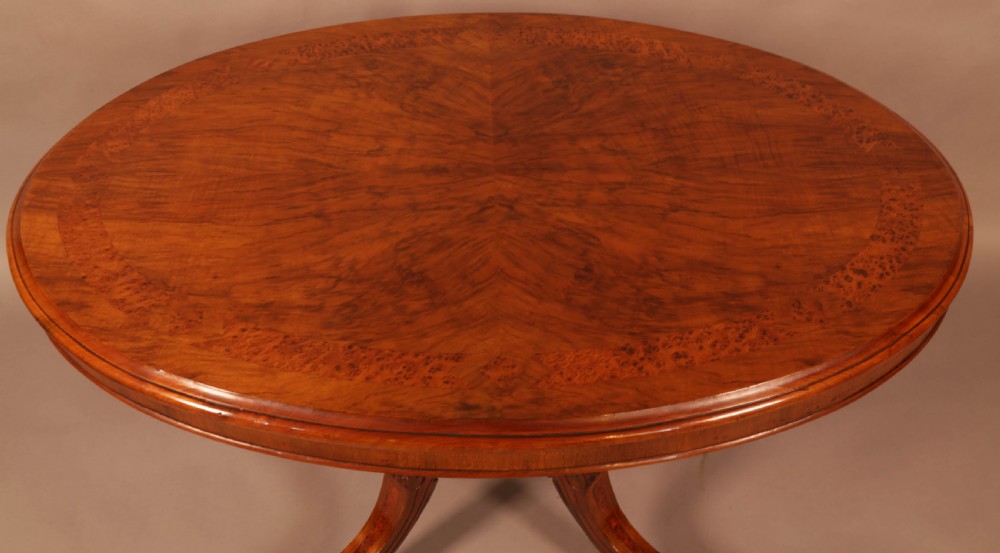 An oval Victorian burr walnut table featuring a figured veneer top and a central carved pedestal with four scroll feet. View V