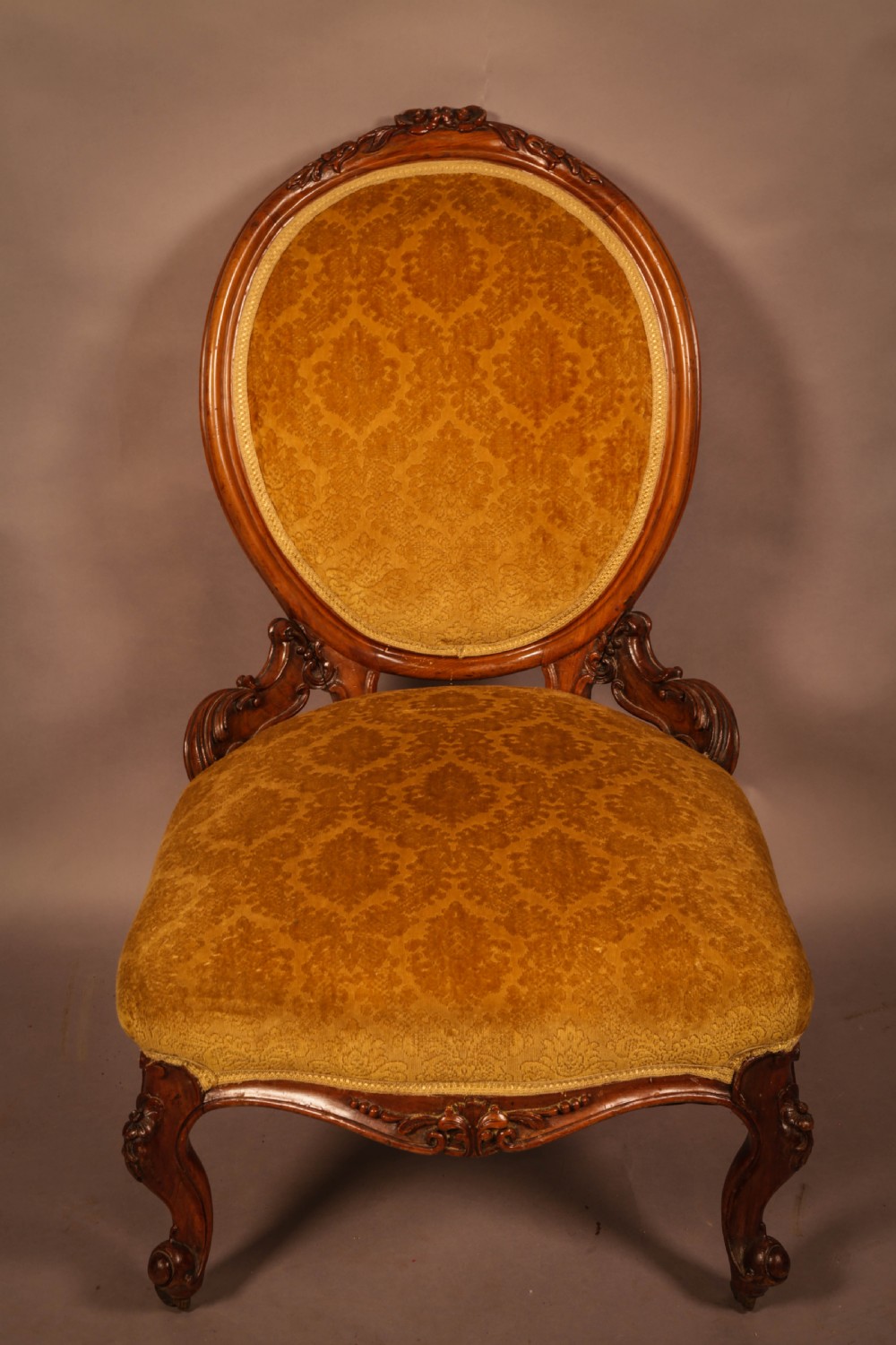 A Victorian walnut nursing chair featuring carved framework, an oval backrest, and golden patterned upholstery.