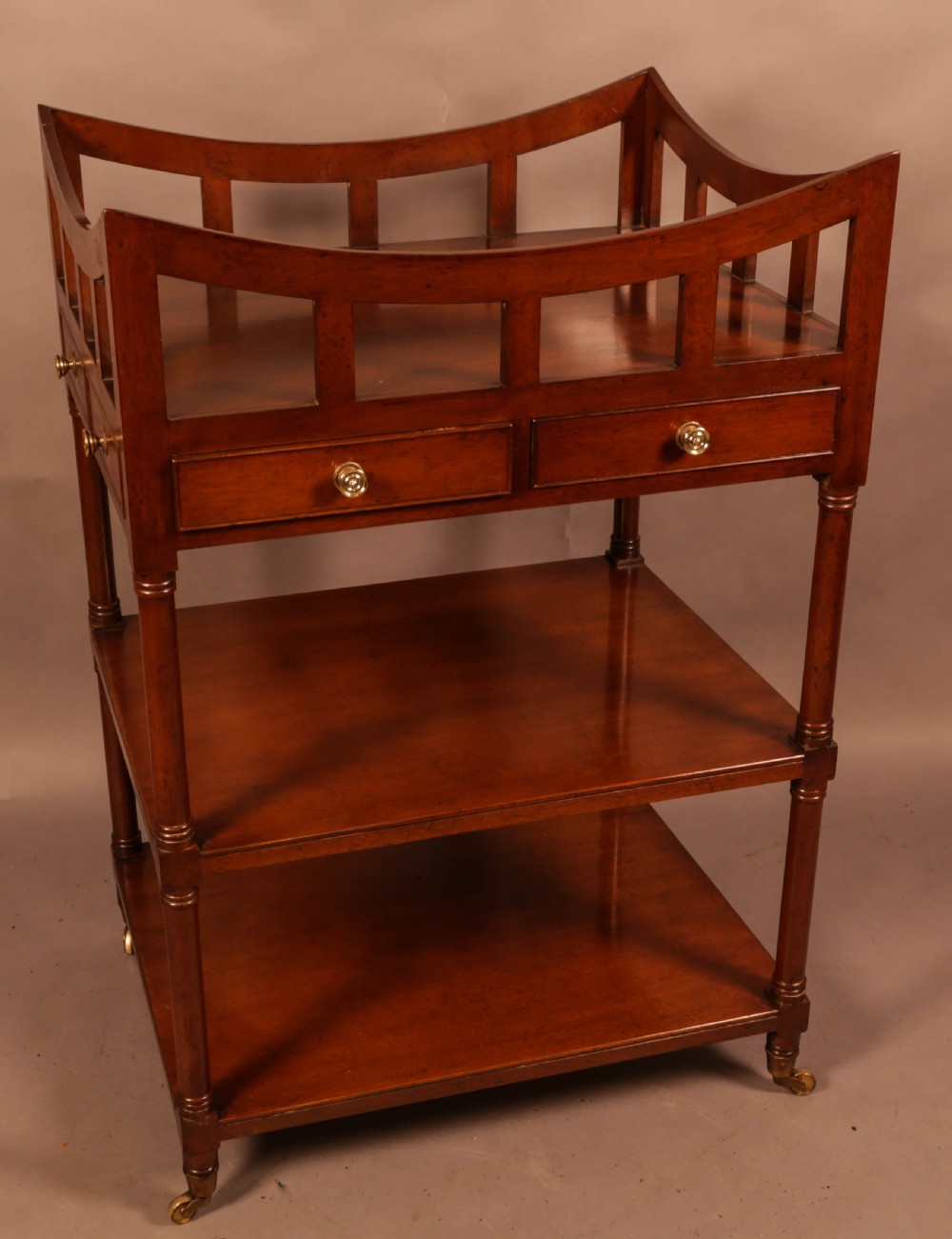A three-tier mahogany library book stand featuring open shelves, two small drawers with brass knobs, and turned supports on castors.