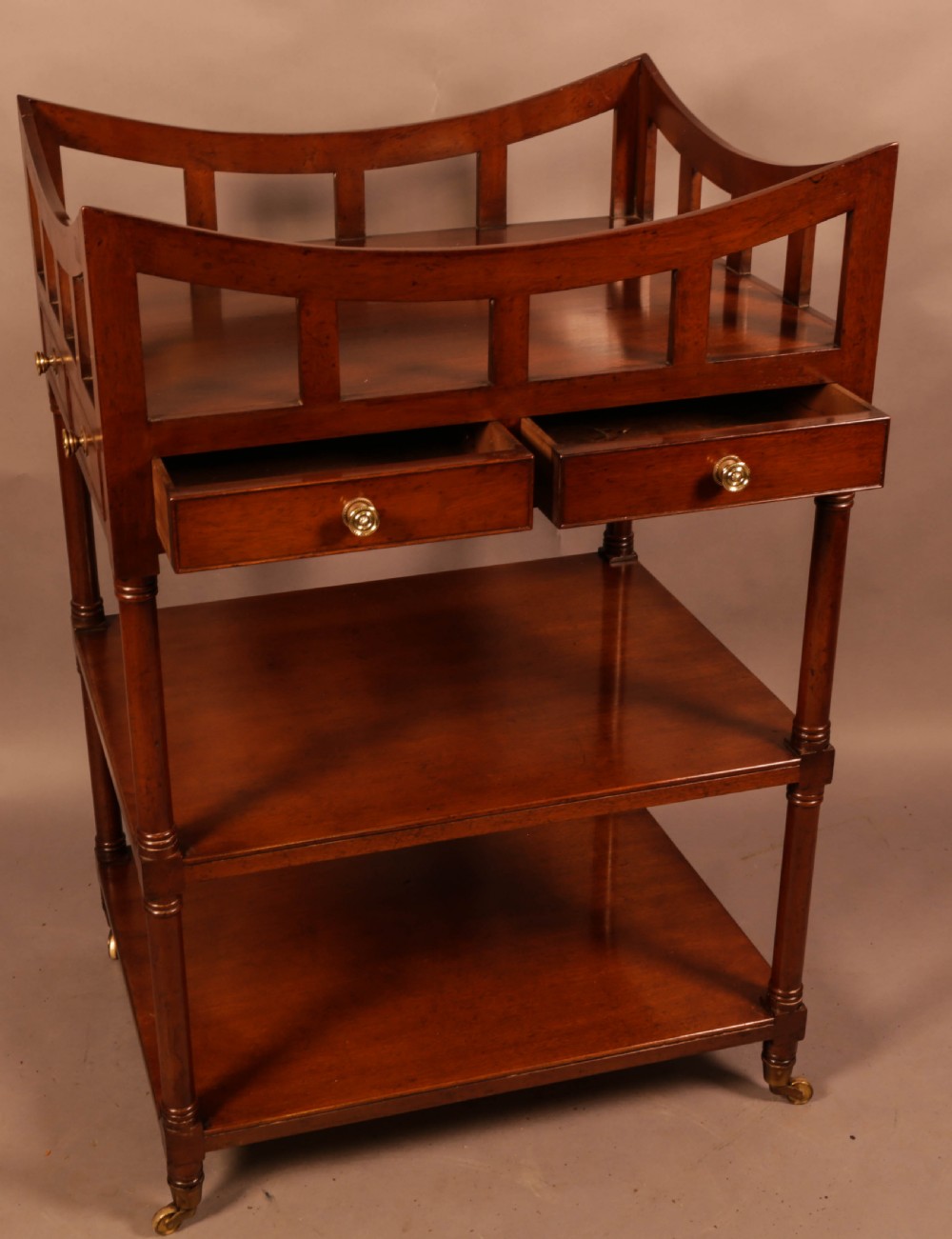 A three-tier mahogany library book stand featuring open shelves, two small drawers with brass knobs, and turned supports on castors. View II