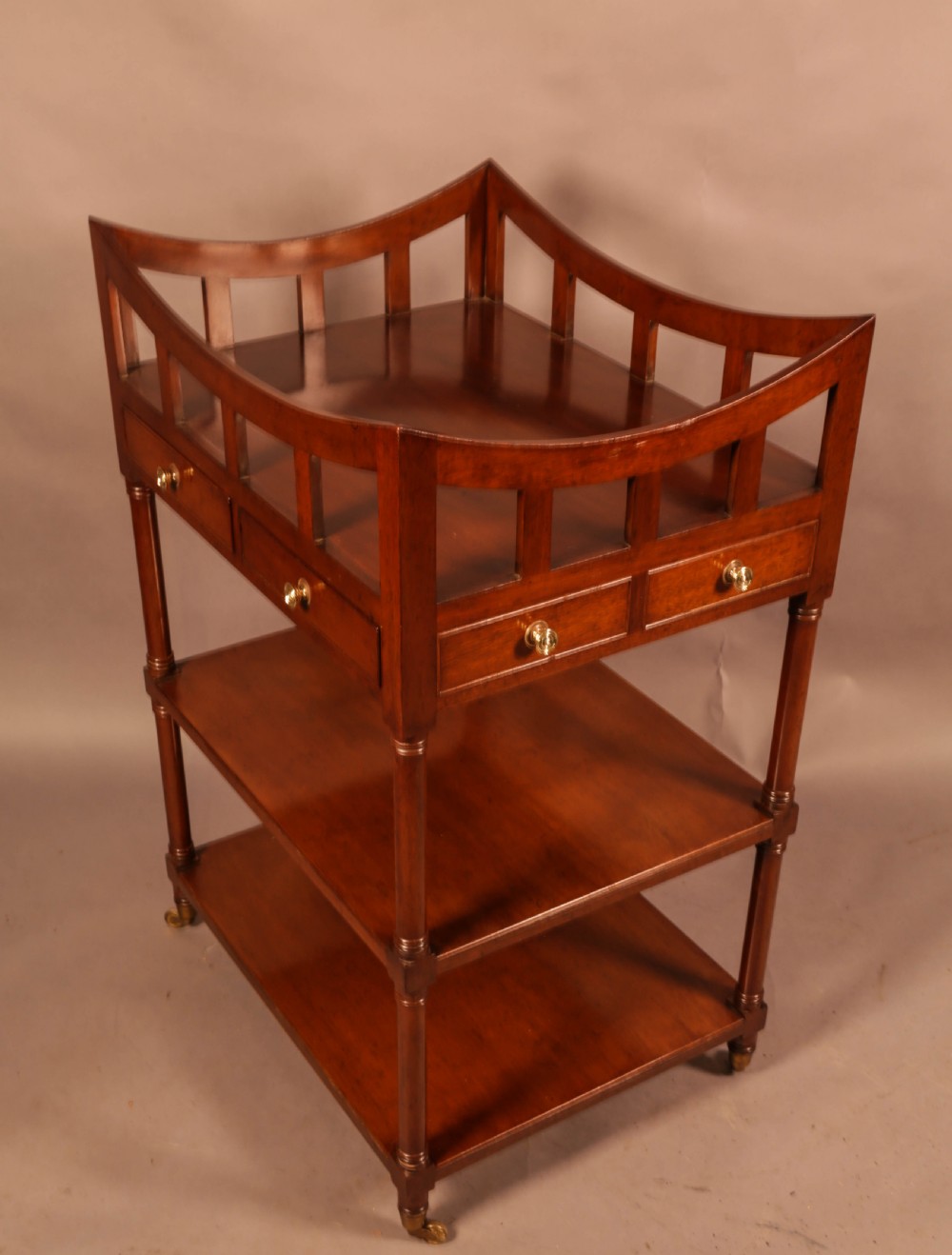 A three-tier mahogany library book stand featuring open shelves, two small drawers with brass knobs, and turned supports on castors. View IV