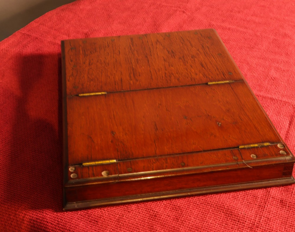 An angled shot of an antique wooden campaign mirror resting on a red textured surface, showing its folding frame and built-in base compartment. View V