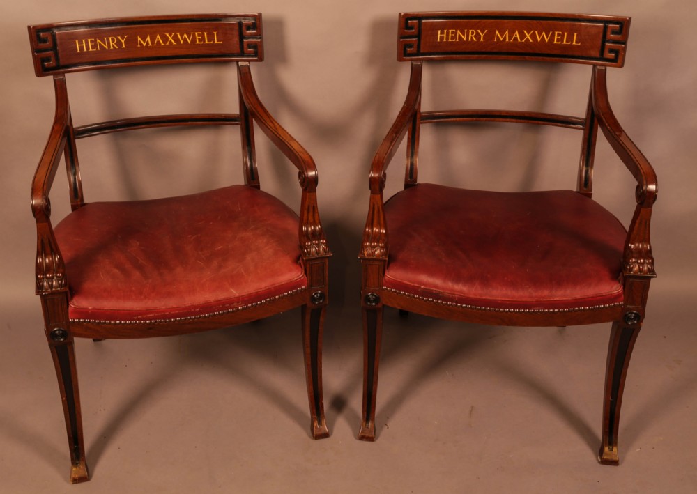 A pair of Regency mahogany carver chairs featuring red leather seats, sabre legs, ebony Greek key inlay, and the name Henry Maxwell gilt-stamped across the top rails.