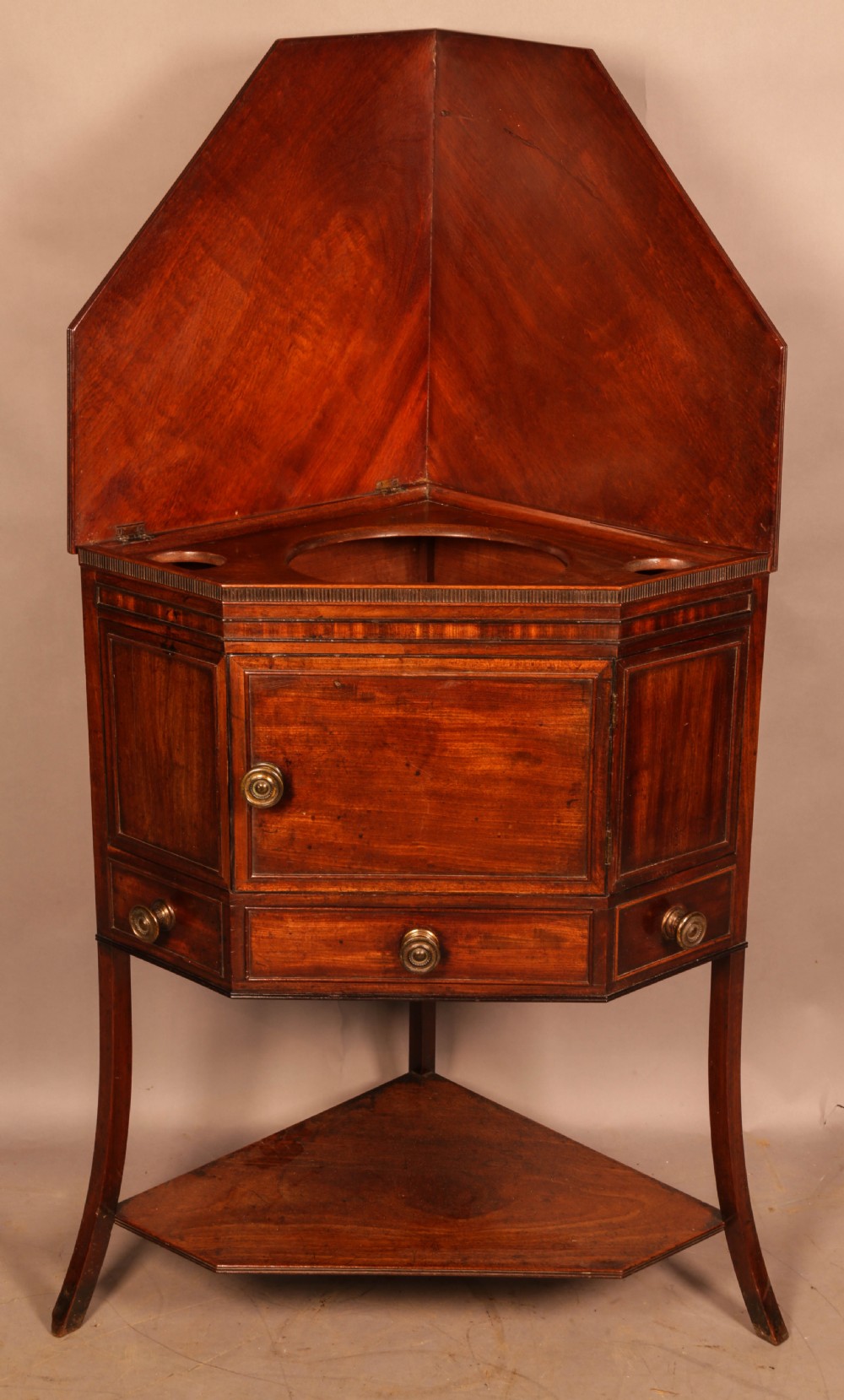 An 18th-century Georgian mahogany corner washstand featuring a hinged splashback, central basin opening, single cupboard door, brass knob handles, and a lower undertier shelf.