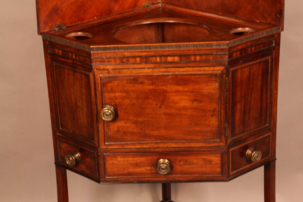 An 18th-century Georgian mahogany corner washstand featuring a hinged splashback, central basin opening, single cupboard door, brass knob handles, and a lower undertier shelf. View VI