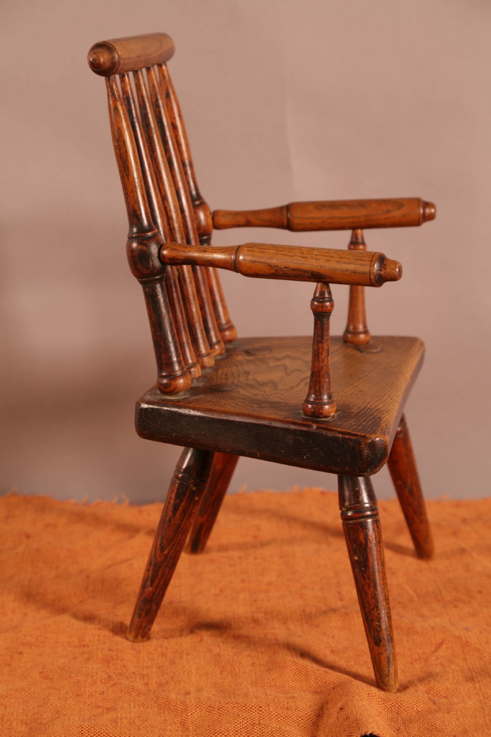 An antique wooden childrens spindle-back armchair featuring a solid saddle seat, turned armrests, and splayed legs with a warm brown patinated finish. View II