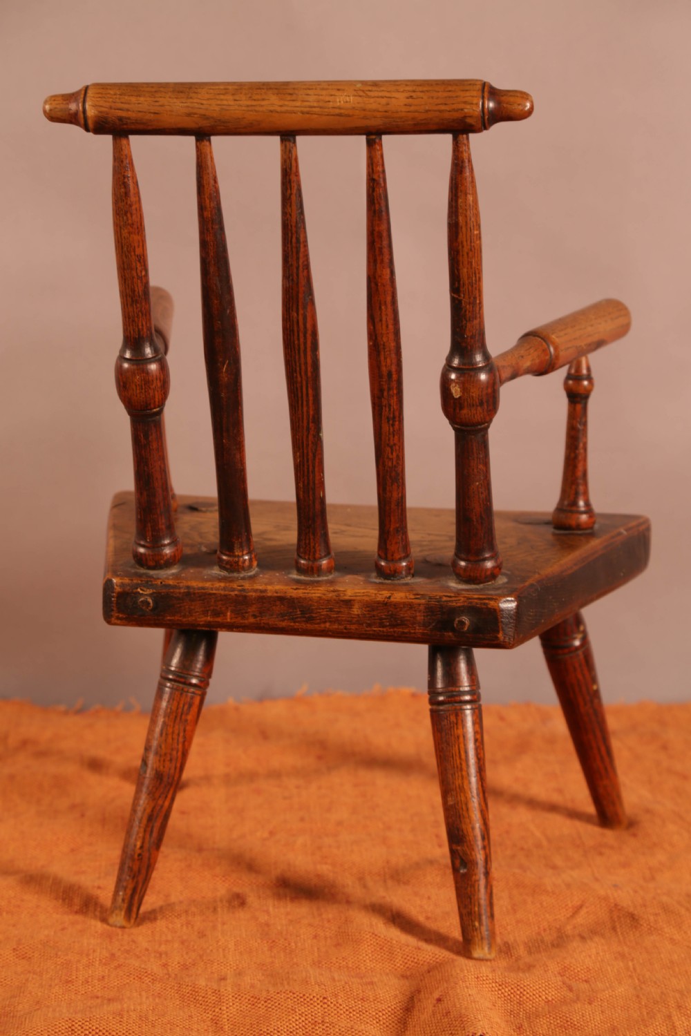 An antique wooden childrens spindle-back armchair featuring a solid saddle seat, turned armrests, and splayed legs with a warm brown patinated finish. View III