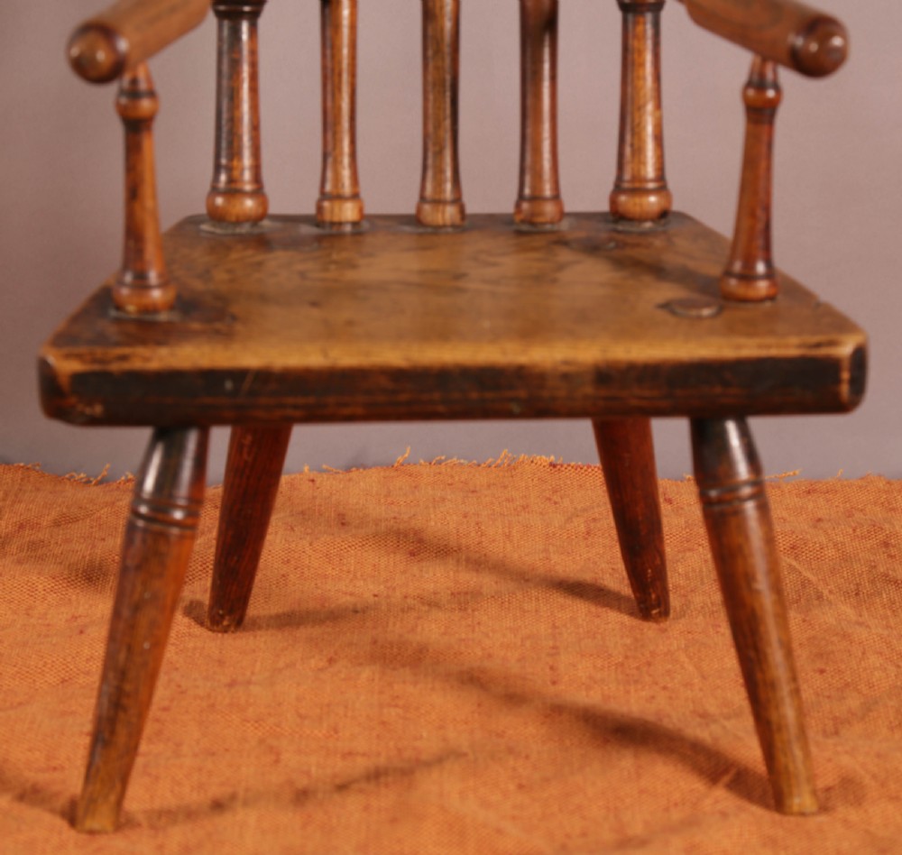 An antique wooden childrens spindle-back armchair featuring a solid saddle seat, turned armrests, and splayed legs with a warm brown patinated finish. View V