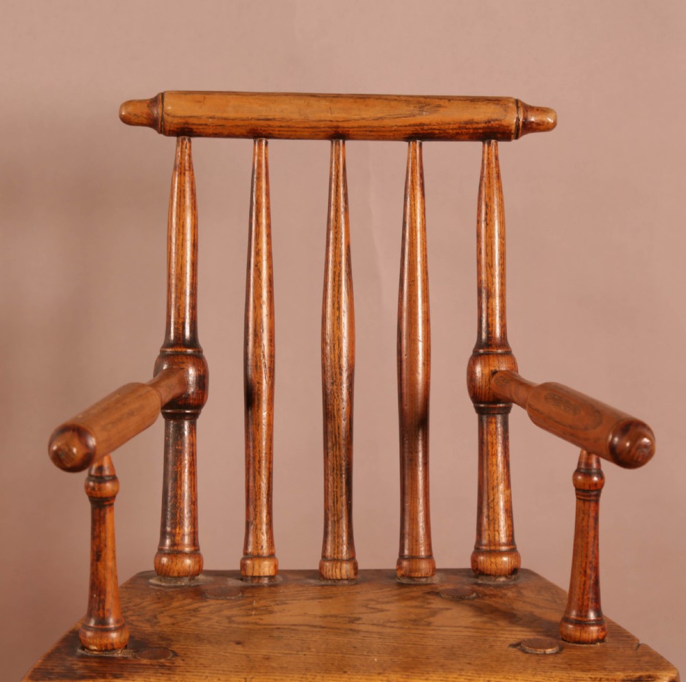 An antique wooden childrens spindle-back armchair featuring a solid saddle seat, turned armrests, and splayed legs with a warm brown patinated finish. View VI