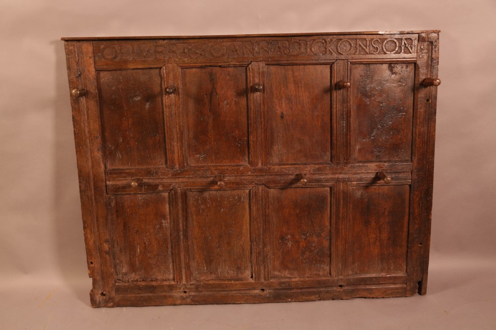 A 17th-century carved dark oak wall panel featuring an inscribed top frieze reading Oliver and Joanna Dickonson, framing double-tier field panels with wooden peg hooks.