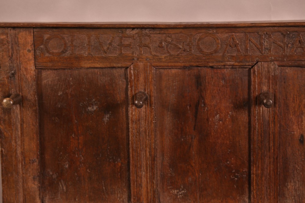 A 17th-century carved dark oak wall panel featuring an inscribed top frieze reading Oliver and Joanna Dickonson, framing double-tier field panels with wooden peg hooks. View II