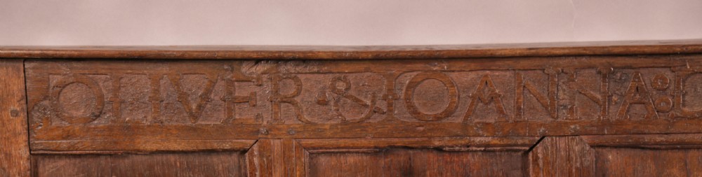 A 17th-century carved dark oak wall panel featuring an inscribed top frieze reading Oliver and Joanna Dickonson, framing double-tier field panels with wooden peg hooks. View III