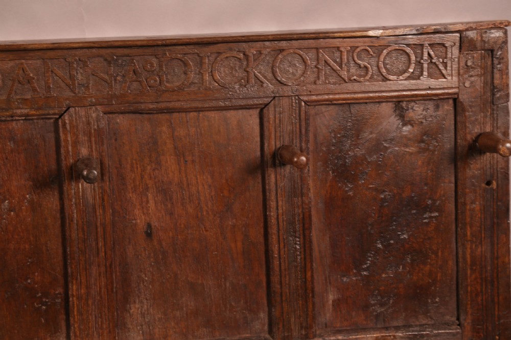 A 17th-century carved dark oak wall panel featuring an inscribed top frieze reading Oliver and Joanna Dickonson, framing double-tier field panels with wooden peg hooks. View IV