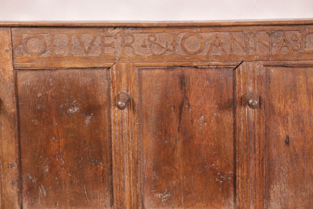 A 17th-century carved dark oak wall panel featuring an inscribed top frieze reading Oliver and Joanna Dickonson, framing double-tier field panels with wooden peg hooks. View V