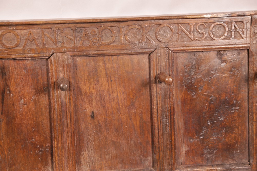 A 17th-century carved dark oak wall panel featuring an inscribed top frieze reading Oliver and Joanna Dickonson, framing double-tier field panels with wooden peg hooks. View VI