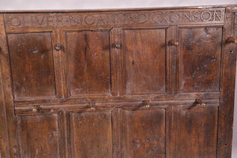 A 17th-century carved dark oak wall panel featuring an inscribed top frieze reading Oliver and Joanna Dickonson, framing double-tier field panels with wooden peg hooks. View VII