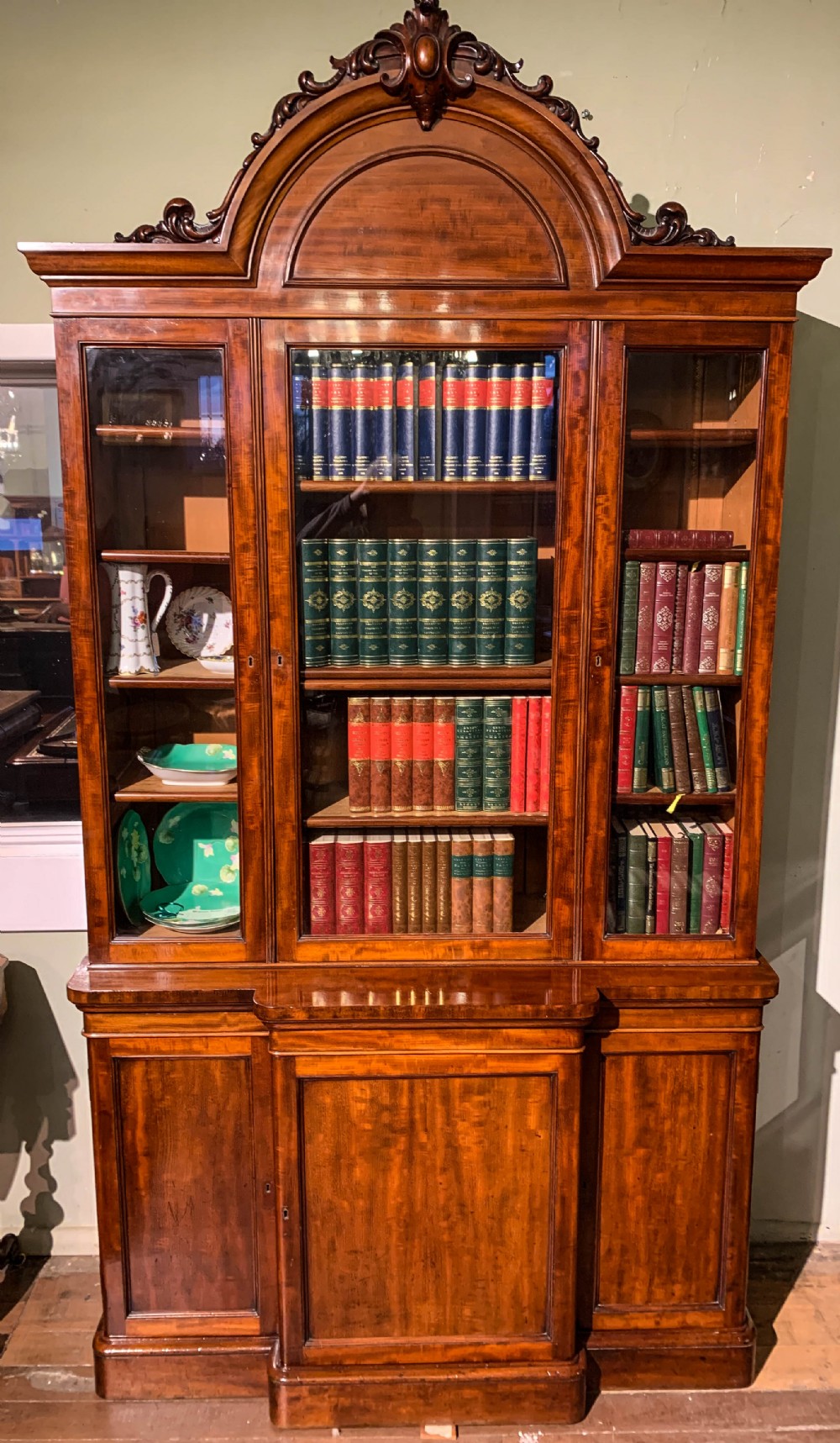 A 19th-century mahogany library bookcase featuring a carved arch pediment, glazed upper doors filled with books, and panelled cupboard doors below.