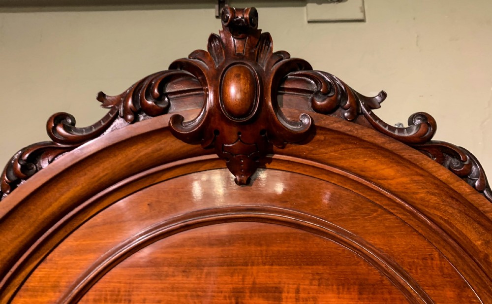 A 19th-century mahogany library bookcase featuring a carved arch pediment, glazed upper doors filled with books, and panelled cupboard doors below. View III