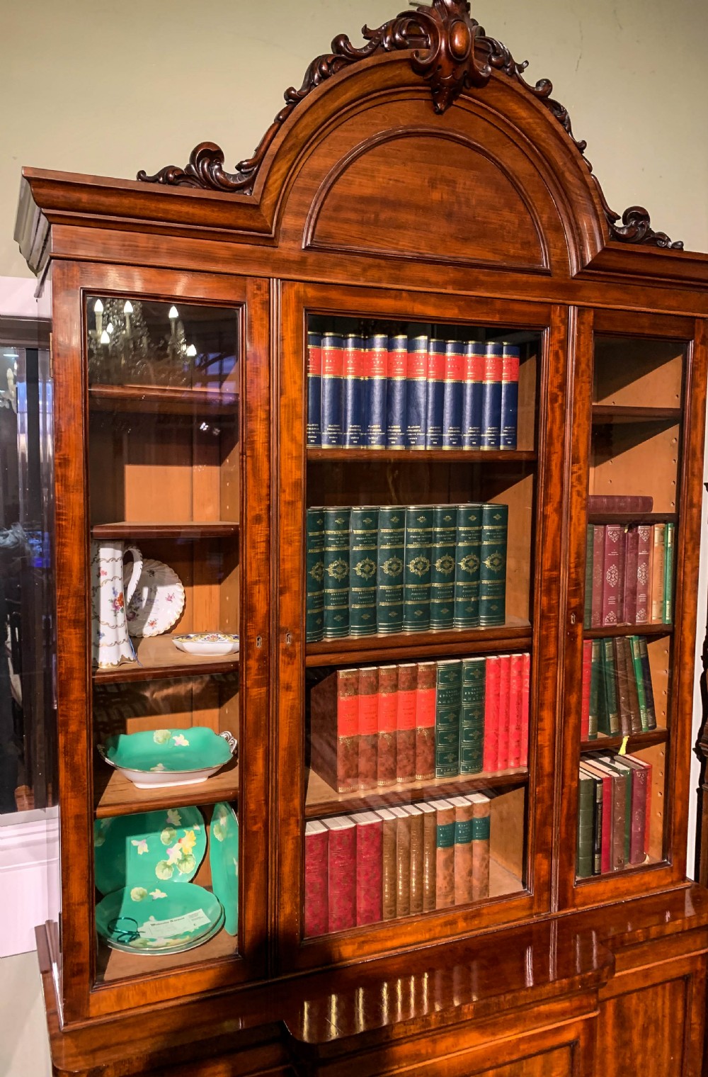 A 19th-century mahogany library bookcase featuring a carved arch pediment, glazed upper doors filled with books, and panelled cupboard doors below. View V