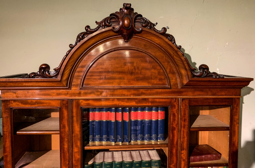 A 19th-century mahogany library bookcase featuring a carved arch pediment, glazed upper doors filled with books, and panelled cupboard doors below. View VI
