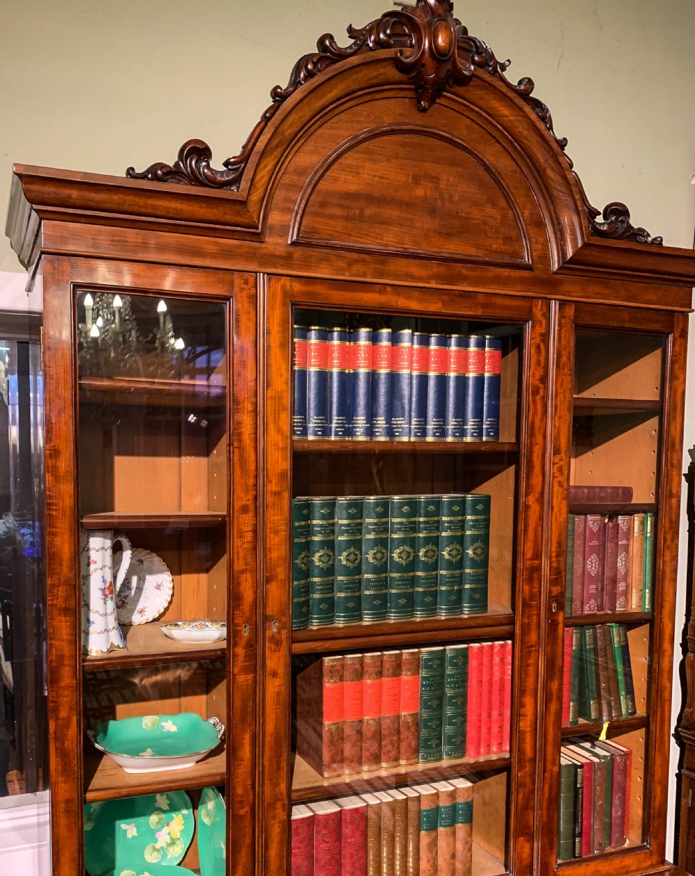 A 19th-century mahogany library bookcase featuring a carved arch pediment, glazed upper doors filled with books, and panelled cupboard doors below. View VIII