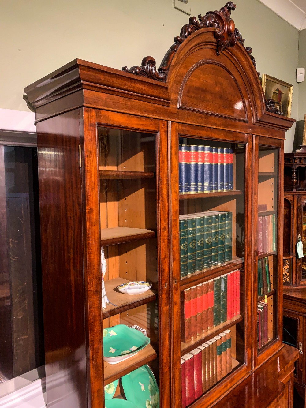 A 19th-century mahogany library bookcase featuring a carved arch pediment, glazed upper doors filled with books, and panelled cupboard doors below. View X
