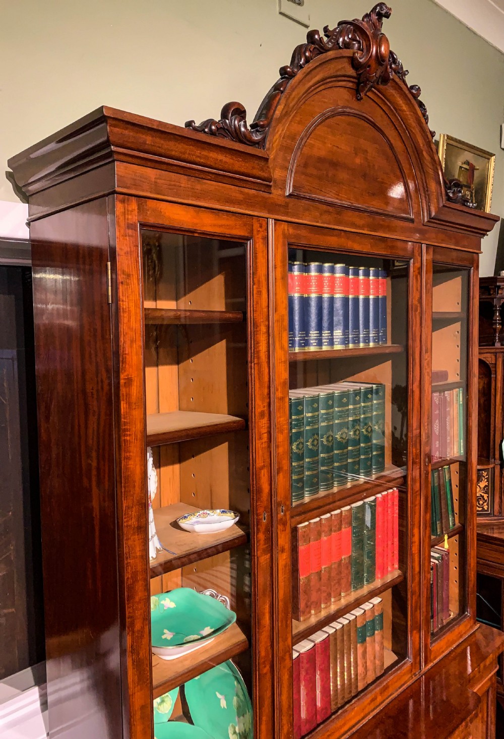 A 19th-century mahogany library bookcase featuring a carved arch pediment, glazed upper doors filled with books, and panelled cupboard doors below. View XII