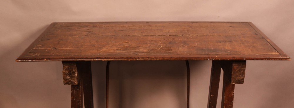 An antique dark timber trestle side table featuring a plank top supported by splayed legs and curved wrought iron stretchers. View VII